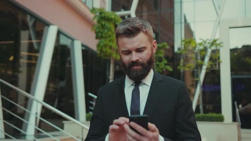 Serious Successful Business Man Uses Smartphone While Walking on the Big City Business District