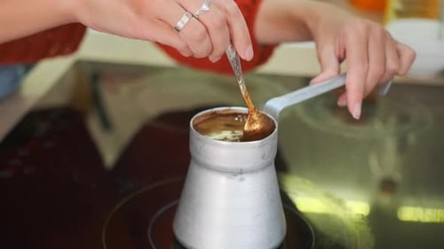 Weibliche Hände in rotem Pullover rühren hausgemachten Kaffee in einer Cezve auf einem Elektroherd um