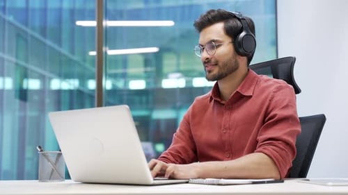 A focused IT specialist in wireless headphones works on a laptop sitting at a workplace