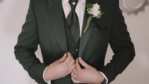 Man in a dark green suit adjusting his jacket, wearing a boutonniere, preparing for an event.