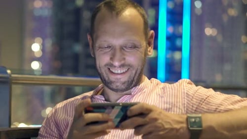 Young, Happy Man Playing Game on Smartphone Sitting on Terrace at Night 30s