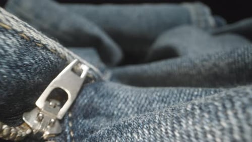 The camera glides across blue denim pants, passing through a metallic zipper. Texture. Dolly slider