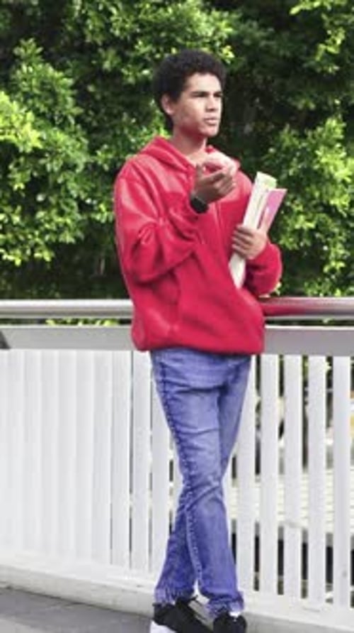 Vertical video: Leaning teen in hoodie holding notes and apple laughing as peer entering on balcony