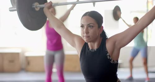 Determined unaltered biracial woman free weight training at gym lifting barbell, in slow motion