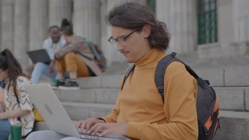 Student Sitting on Stairs Outside the University and Typing on Laptop
