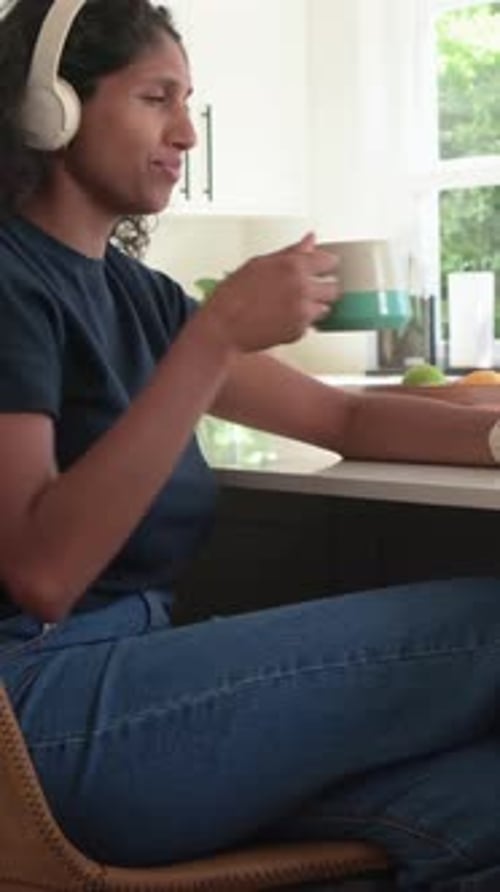 Vertical video: Smiling woman in navy tee headset reaching for mug, sipping for break in kitchen