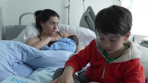 Mother breastfeeding newborn while older sibling in red jacket sits nearby on hospital bed.
