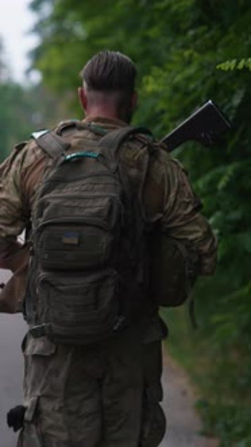 A Soldier Armed with a Weapon and Carrying a Backpack is Walking on the Road