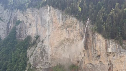 Wide aerial of waterfall on rocky mountainside with forest on top