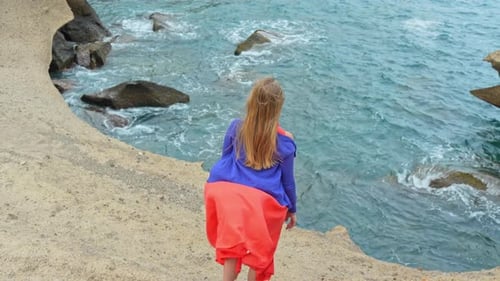 Wide shot of woman raising arms facing ocean waves, windy day, tilt up, day