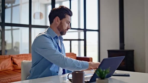 Freelancer barbudo abrindo a tampa do laptop sentado ao lado do local de trabalho do apartamento
