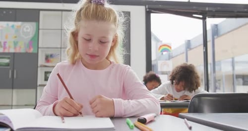 Video of happy caucasian schoolgirl sitting at desk writing in diverse school class