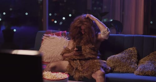 Stone Age Human in Anima Fur Sitting on the Sofa Using Mobile Phone Like Real Modern Person Staying
