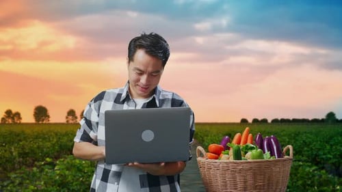 Asiatischer männlicher Bauer mit Gemüsekorb, der lächelt, während er den Laptop auf dem Feld benutzt