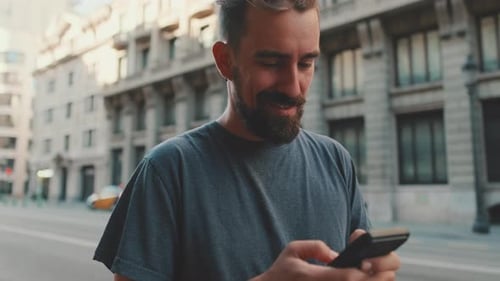 Young smiling man with beard stands on street using cellphone
