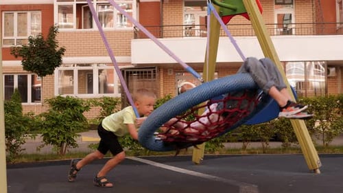 A Boy Rides a Girl on a Round Swing with a Net in the Yard on a Playground