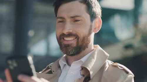 Face Smiling Stylish Handsome Man with Beard in Coat Uses Smartphone Sitting in Part at Sunlight
