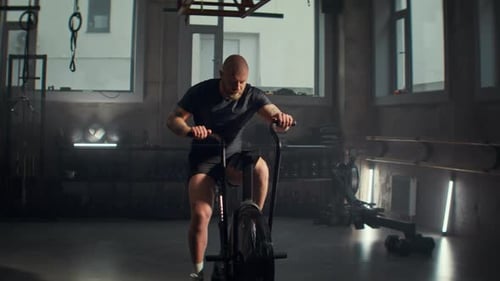 Dedicated Man in Tracksuit Exercises on Air Bike Trainer in Gym