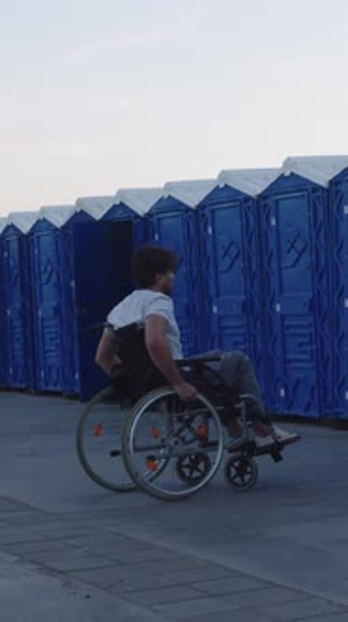 Man in Wheelchair Rides Along Row of Portable Toilets