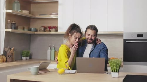 Smiling Couple Embracing While Using Laptop in Kitchen