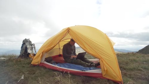 Um homem trabalha em um computador enquanto está sentado em uma barraca com uma paisagem montanhosa