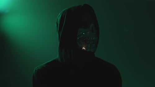 Hacker encapuzado com máscara facial olhando para a câmera no quarto escuro