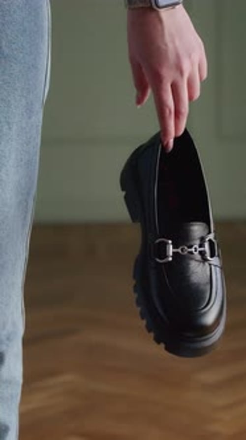 Hand Pointing at a Black Loafer on Wooden Floor with a Partial View of a Person Wearing Jeans