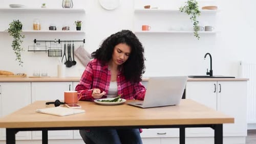 Woman Eats Salad While Working on Laptop in Kitchen