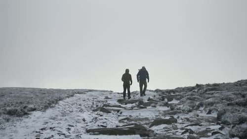Two people hiking from the horizon over frozen, snowy fields and rocks.