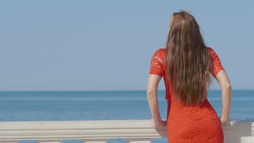 Woman dressed in red near the sea looks at the horizon and enjoys the wind