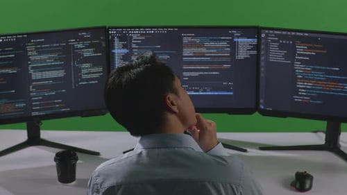 Back View Of Asian Man Thinking While Write Code With Multiple Computer Screens In Green Screen