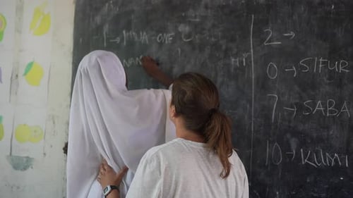 White caucasian teacher helping to black african girl whit white headscarf to write on the blackboar
