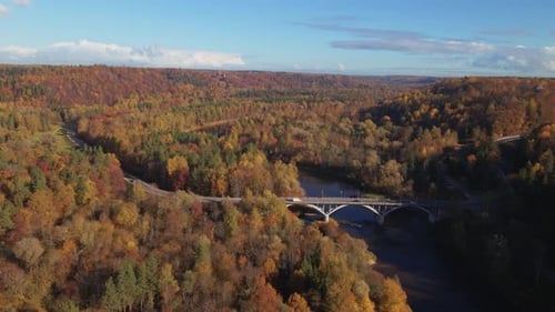 The drone moves right, tracking cars as they cross a bridge over an autumn-colored, hilly landscape,