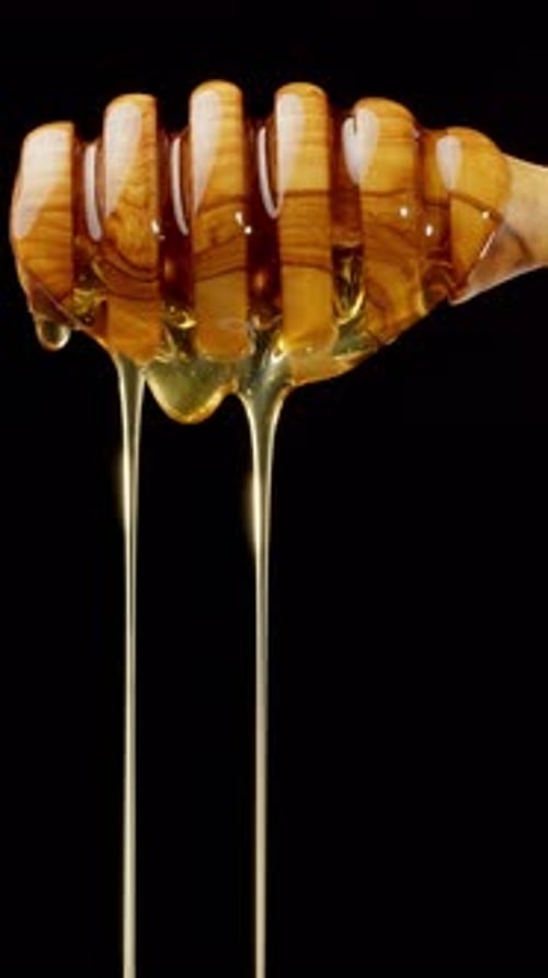 Drops of honey flow from a wooden honey dipper and slowly drip down.