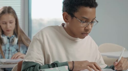 Teenage Left-Handed Boy Sitting at Desk and Writing with Pen at Lesson