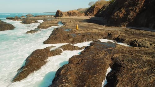Woman in dress standing on volcanic rocky coast surrounded by Pacific Ocean waves