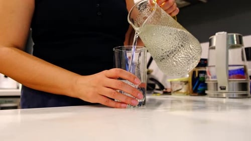 Close Up Shows Hands Pouring Cold Water From a Glass Pitcher Into a Tall Glass on a Kitchen Counter