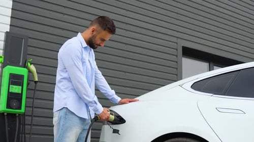 Stylish Man Plugging in Charging Cable to Electric Vehicle and Charges Batteries at Station