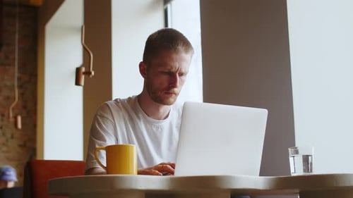 Ein stirnrunzelnder Mann tippt etwas auf der Tastatur seines Laptops Ein Mann in einem weißen T-Shirt arbeitet