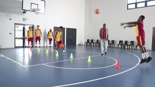 Basketball players practicing dribbling skills in gym with coach observing