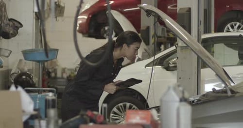 Female car mechanic looking under vehicle bonnet using digital tablet in workshop