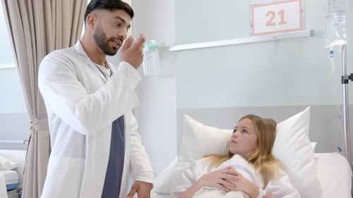 Diverse male doctor showing girl patient in hospital bed how to use asthma inhaler, slow motion