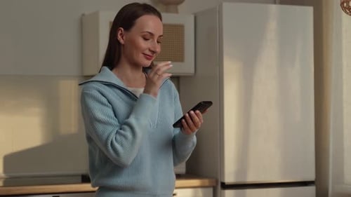 Junge glückliche Hausfrau in einem blauen Pullover mit einem Handy in den Händen auf der Suche nach einem