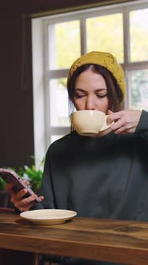 Woman Enjoying Coffee at a Cozy Cafe While Holding a Phone and Wearing a Yellow Beanie in the