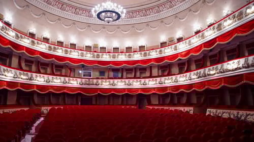 Classic theater indoors. Empty opera house with tiers and red velvet chairs under the large lamp.