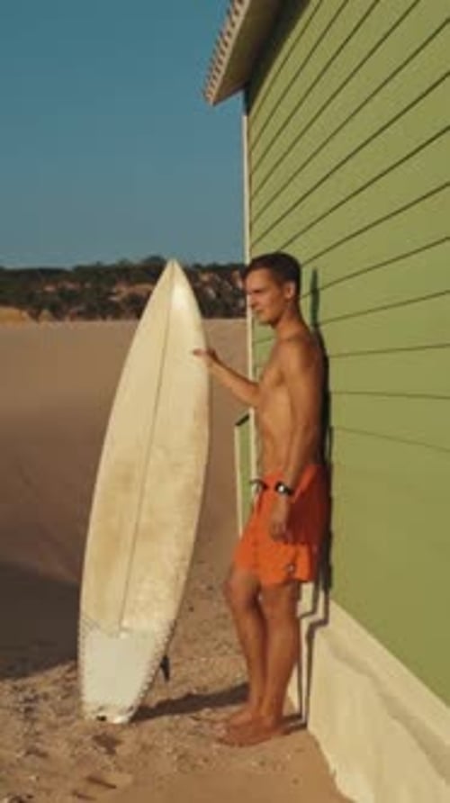 Surfer Leaning on Beach Hut Wall Holding Surfboard