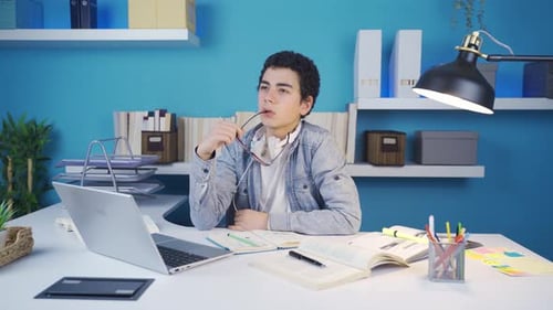 Clever young male student thoughtfully picks up his glasses while looking at laptop.