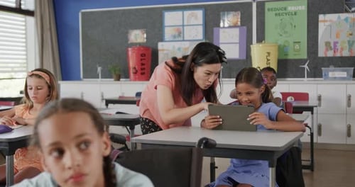 Happy diverse female teacher helping girl using tablet in elementary school class, slow motion