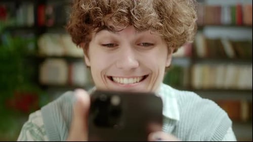 Handsome Curly Student Man Holding Modern Smartphone Texting Message in Home Office or Library