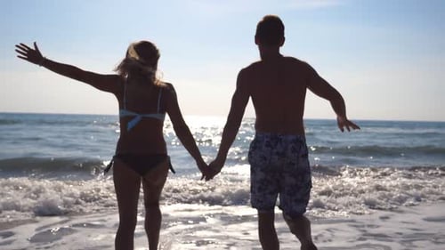 Young Happy Couple Running at the Beach to the Sea and Holding Hands of Each Other Girl and Boy